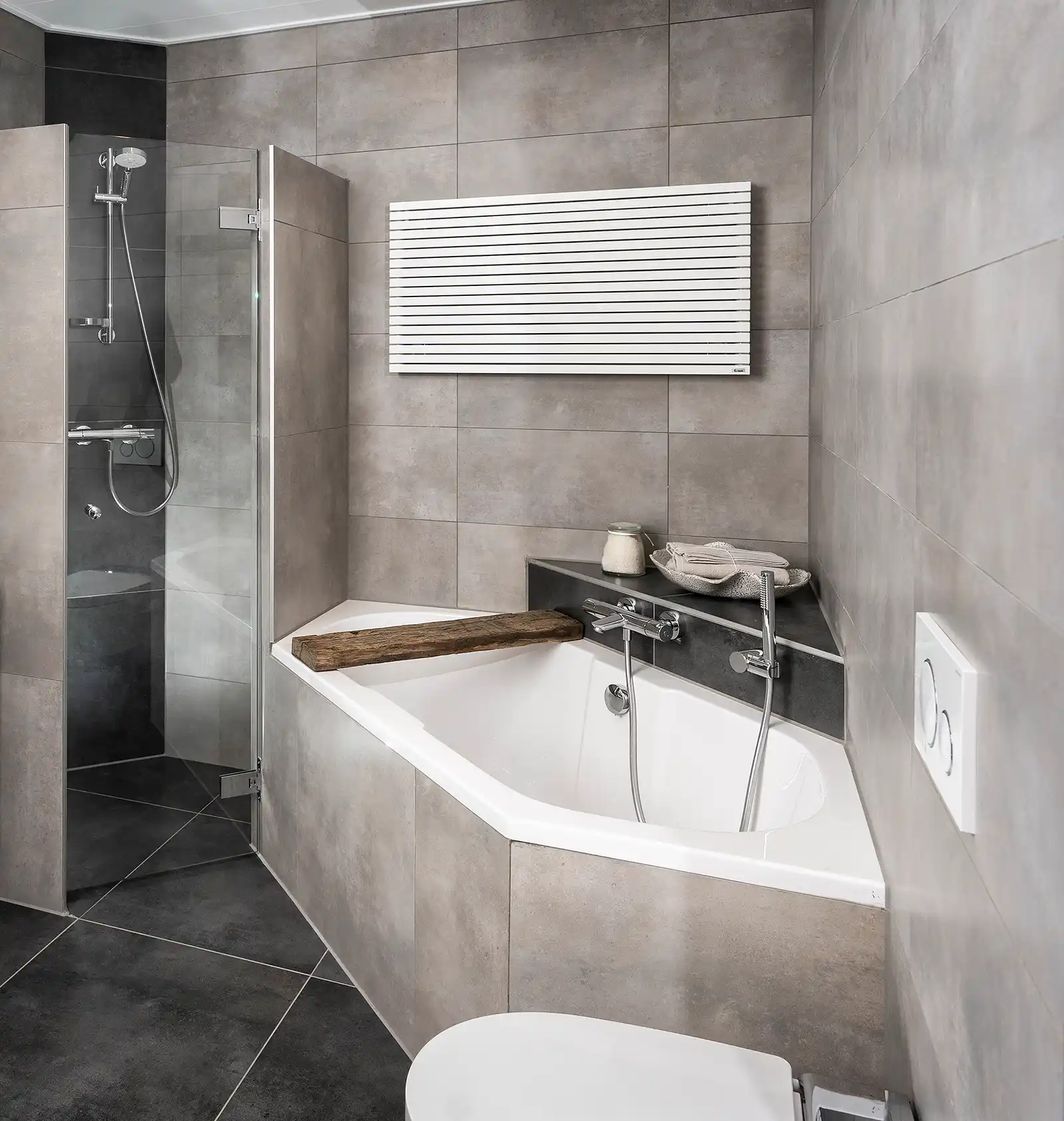 In einem modernen Badezimmer ist eine elegante, schneeweiße Badewanne mit einer Holzablage und chromfarbenen Armaturen sichtbar. Eine transparente Dusche aus Glas ist daneben platziert. Die Wände sind mit grauen Fliesen verkleidet, während ein Heizkörper