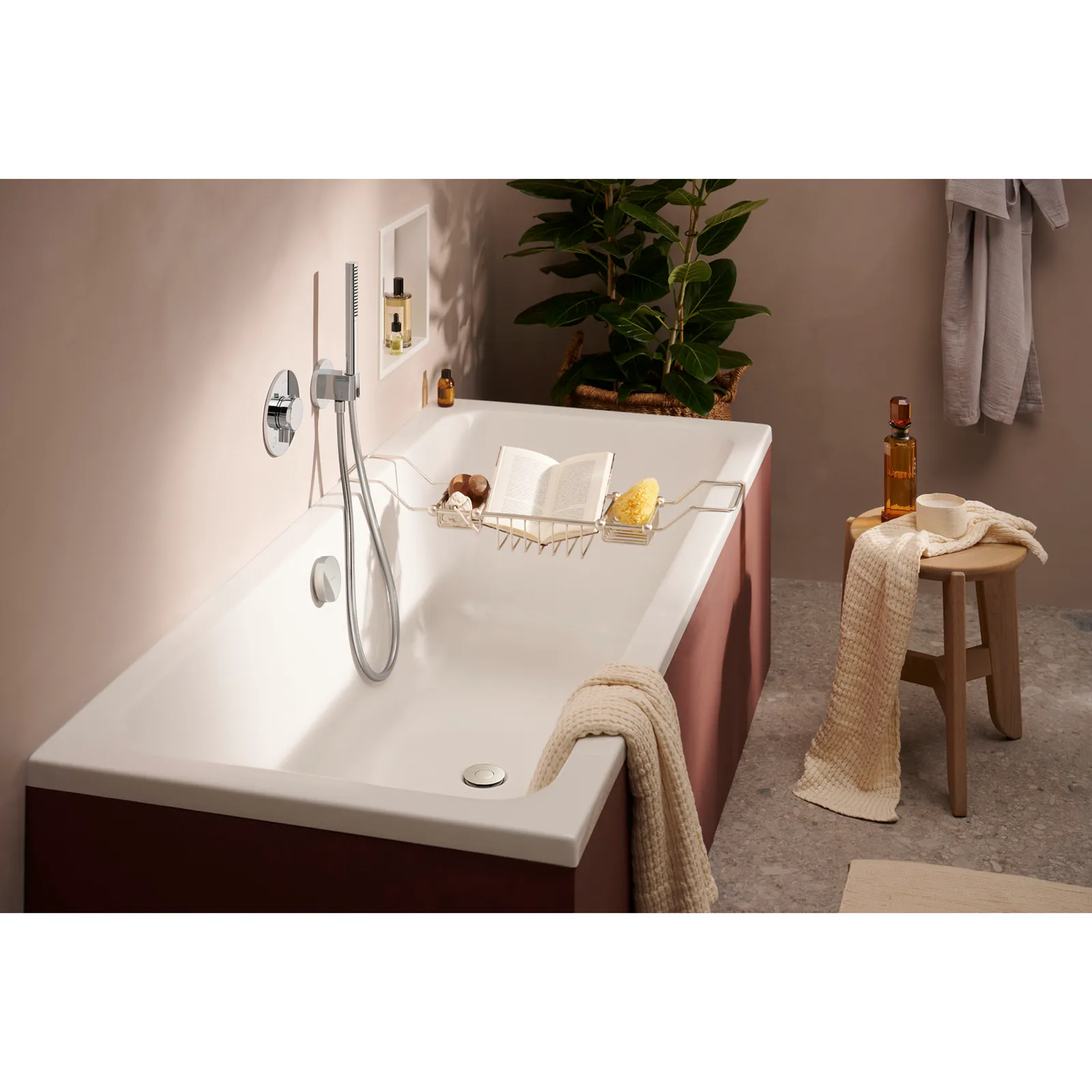Eine weiße Badewanne mit einem eleganten Design, ausgestattet mit einer modernen Armatur. Auf dem Wannenrand liegt ein Buch und Toilettenaccessoires, umgeben von einem gemütlichen Ambiente mit Pflanzen und Dekoration.