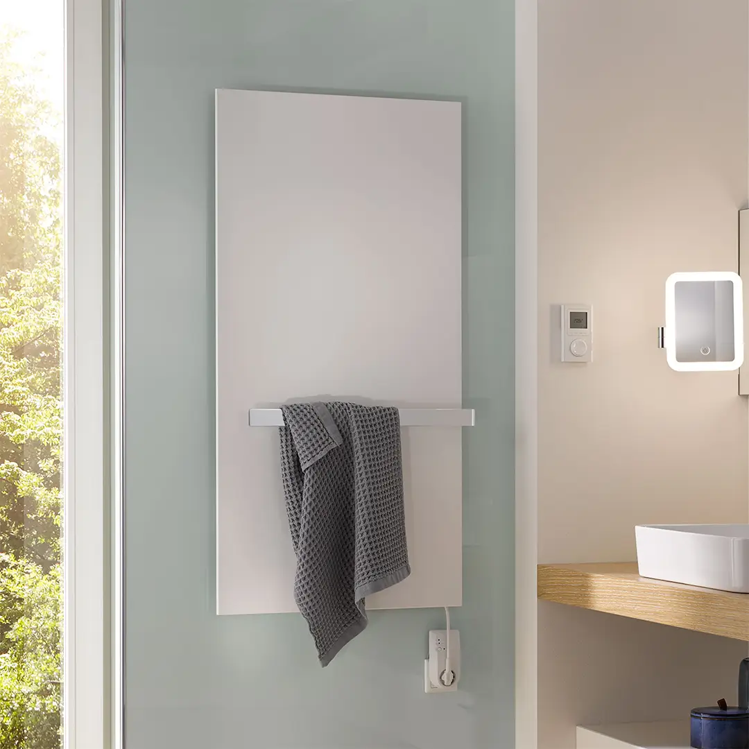Ein großer, vertikaler, weißer Handtuchtrockner hängt an einer modernen Wand. An einem integrierten Halter ist ein graues Handtuch befestigt. Der Bereich ist hell und freundlich, mit einem Blick ins Grüne. Ein kleiner Spiegelschrank ist zu sehen.