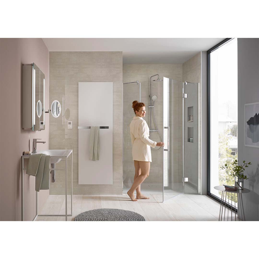 Eine Frau in einem hellen Bademantel steht an einer modernen Duschkabine mit Glaswänden. Der Raum ist hell und minimalistisch gestaltet, mit einem Waschbecken, Spiegeln und einem Fenster, das Natürliches Licht hereinlässt.