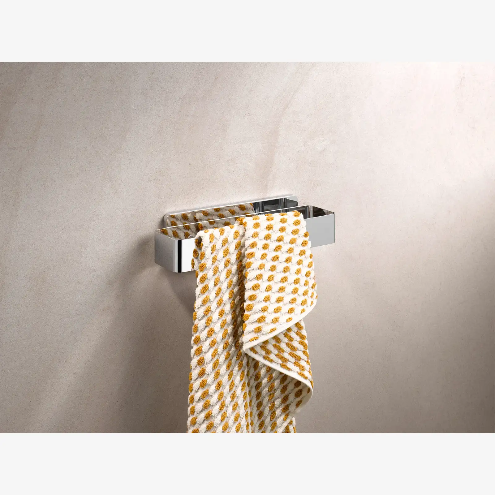 Ein chromfarbener Handtuchhalter ist an einer hellen Wand montiert. Ein Handtuch mit gelb-weißen Mustern hängt über den Halter, der eine moderne, klare Form aufweist.