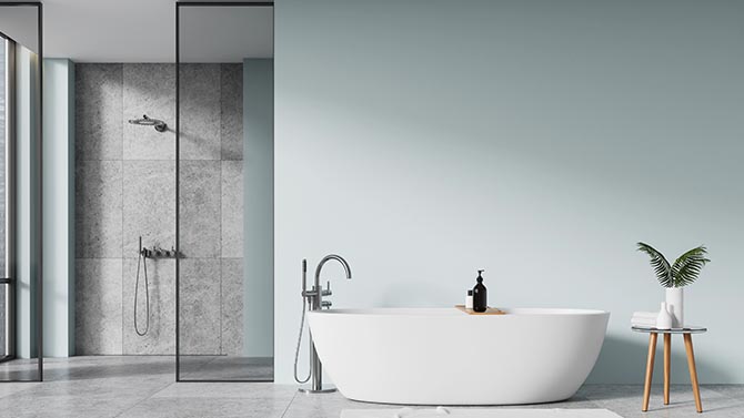 Freistehende weiße Badewanne mit stilvollem Wasserhahn, daneben ein kleiner Tisch mit einer Pflanze und einem Seifenspender. Hintergrund zeigt eine moderne Dusche mit gläserner Trennwand. Wände in sanften Farben.