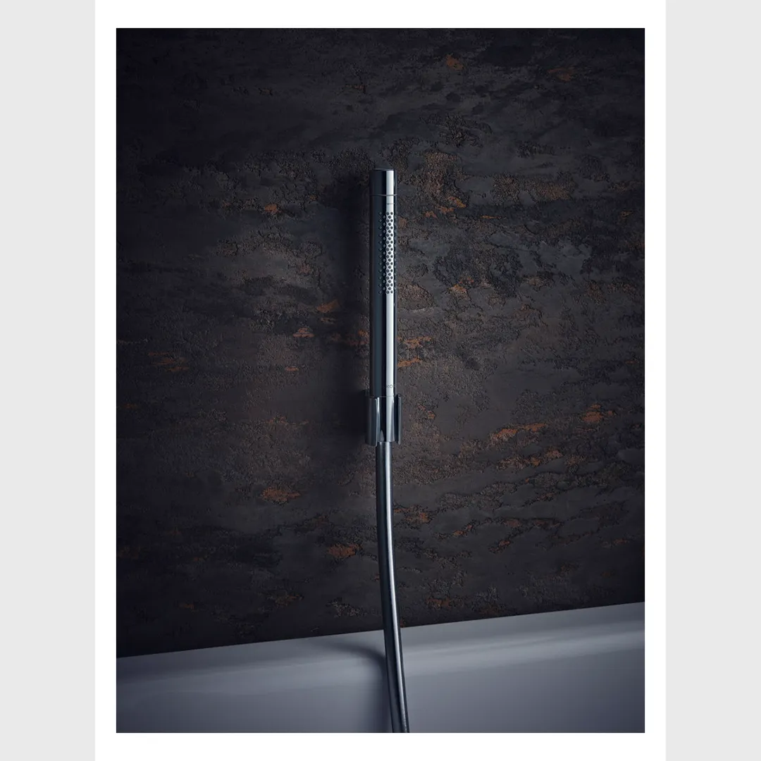 Eine moderne Duschbrause aus verchromtem Metall hängt an einer Wand mit rauer, dunkler Oberfläche. Der Flexschlauch ist sichtbar und die Badewanne ist am unteren Bildrand zu erkennen.