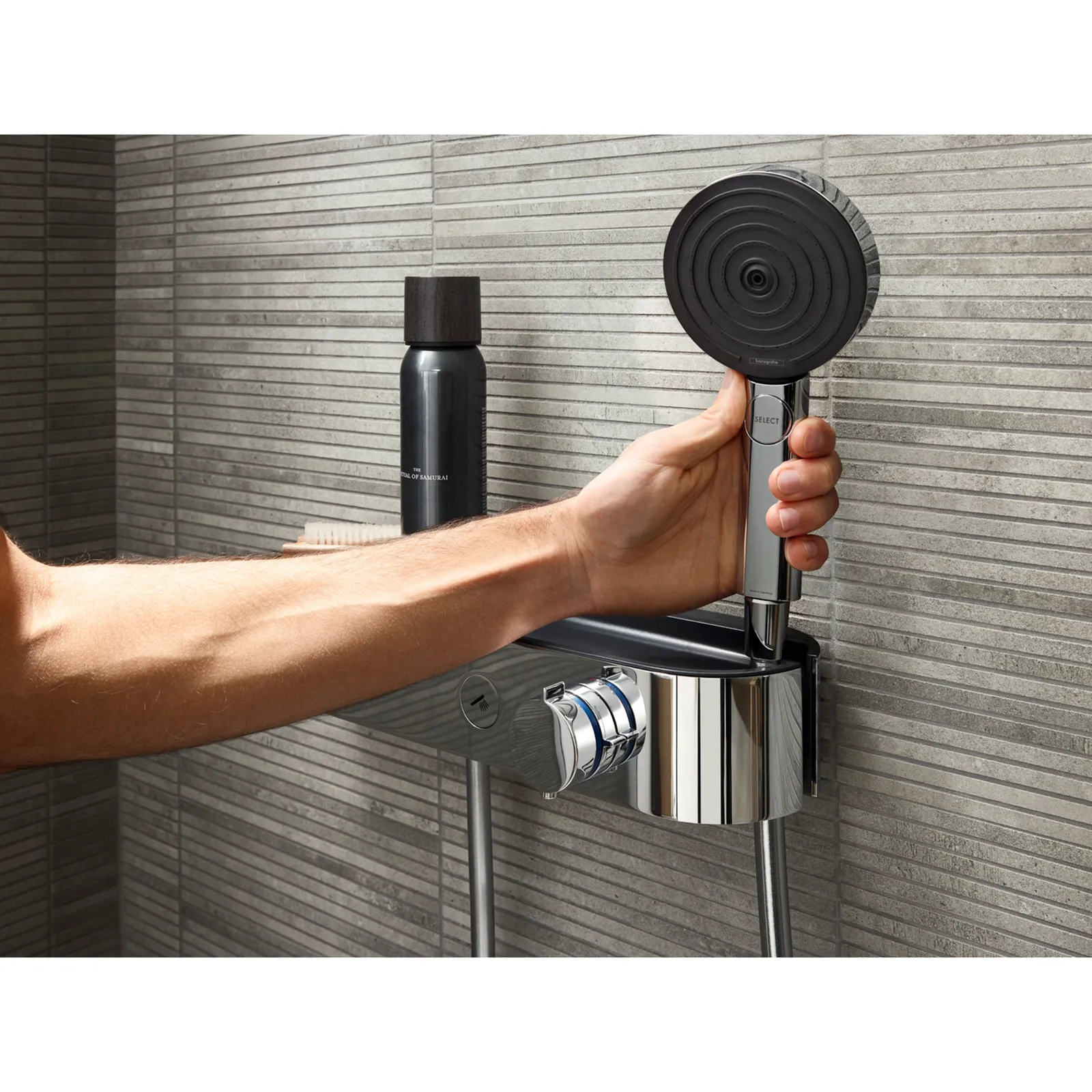 Ein Arm hält einen runden Duschkopf mit einem geschwungenen Design. Die Dusche ist an einer modernen Wand mit strukturierten Fliesen montiert. In der Nähe steht eine schwarze Wasserflasche.