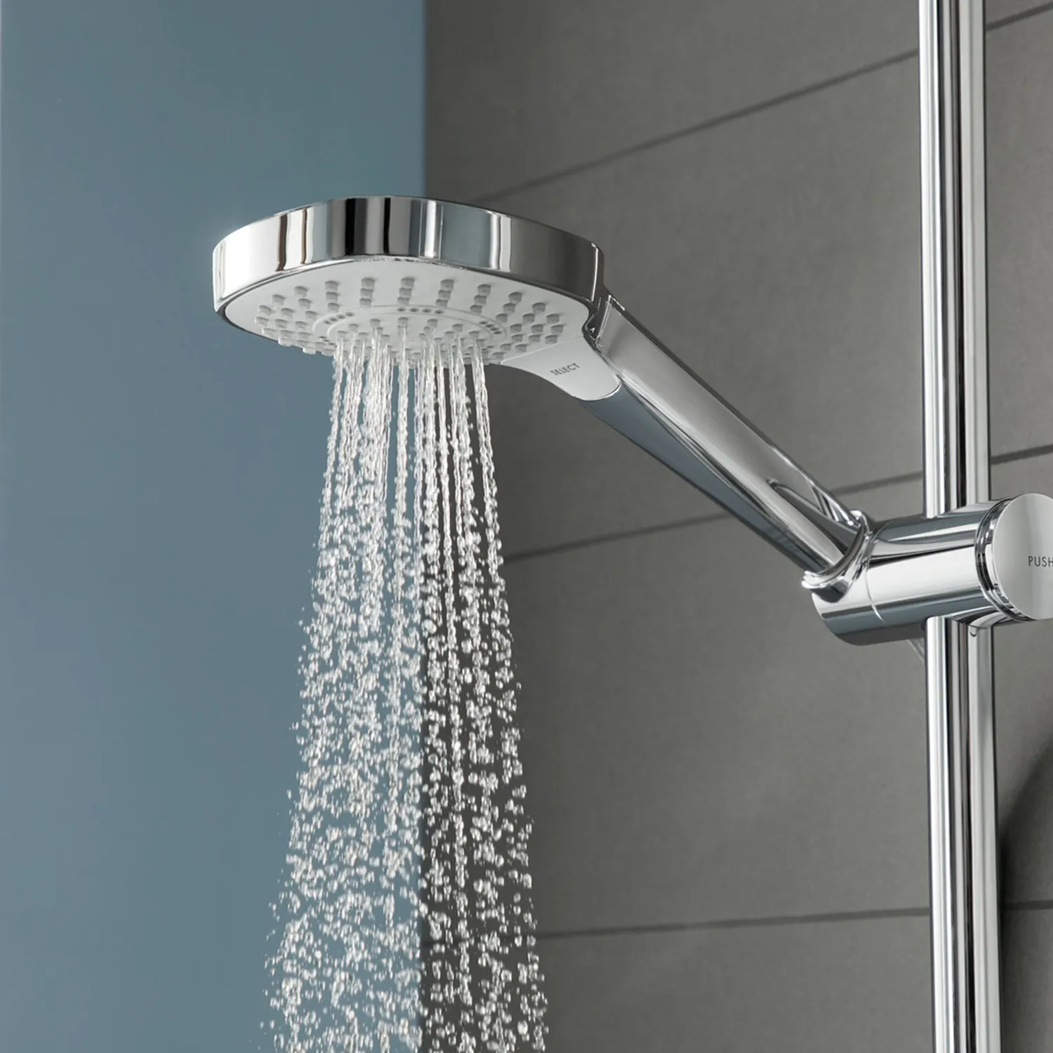 Eine verchromte Handbrause mit flachem, runden Kopf von dem Wasser in feinen Strahlen fließt. Der Hintergrund zeigt eine graue Wandverkleidung, die modern wirkt.