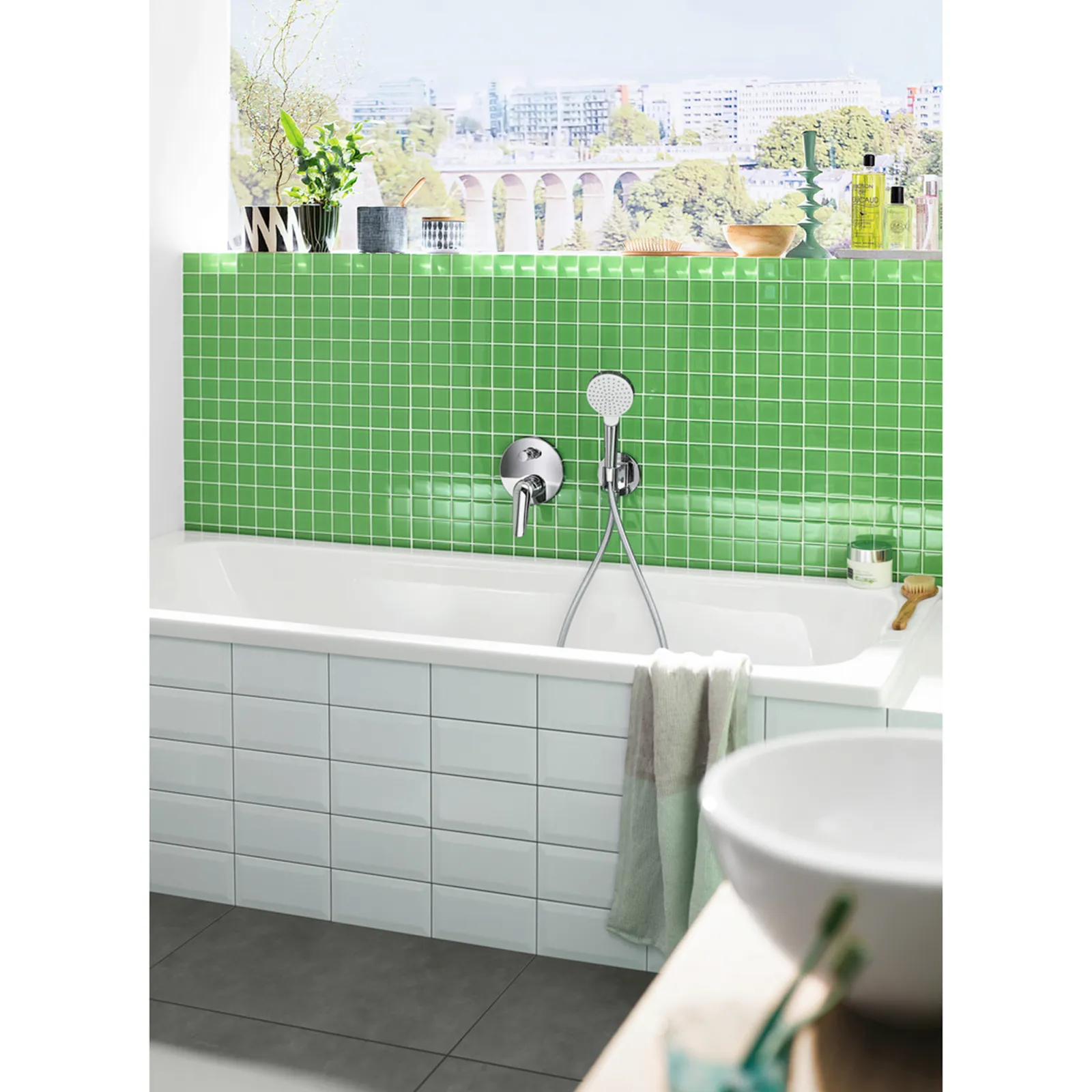 Eine weiße Badewanne ist mit einem glänzenden, verchromten Wasserhahn und einer Handbrause ausgestattet. Die Wand dahinter zeigt grüne Fliesen im Mosaikstil. Das Badezimmer ist hell und modern gestaltet, mit einem großen Fenster im Hintergrund.