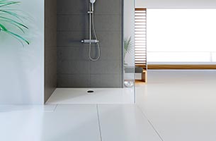Eine moderne Dusche mit einem grauen Fliesenbereich und einer Handbrause ist zu sehen. Der Boden ist weiß und glatt, während im Hintergrund ein heller, skandinavischer Raum mit Holzdetails und großen Fenstern sichtbar ist.