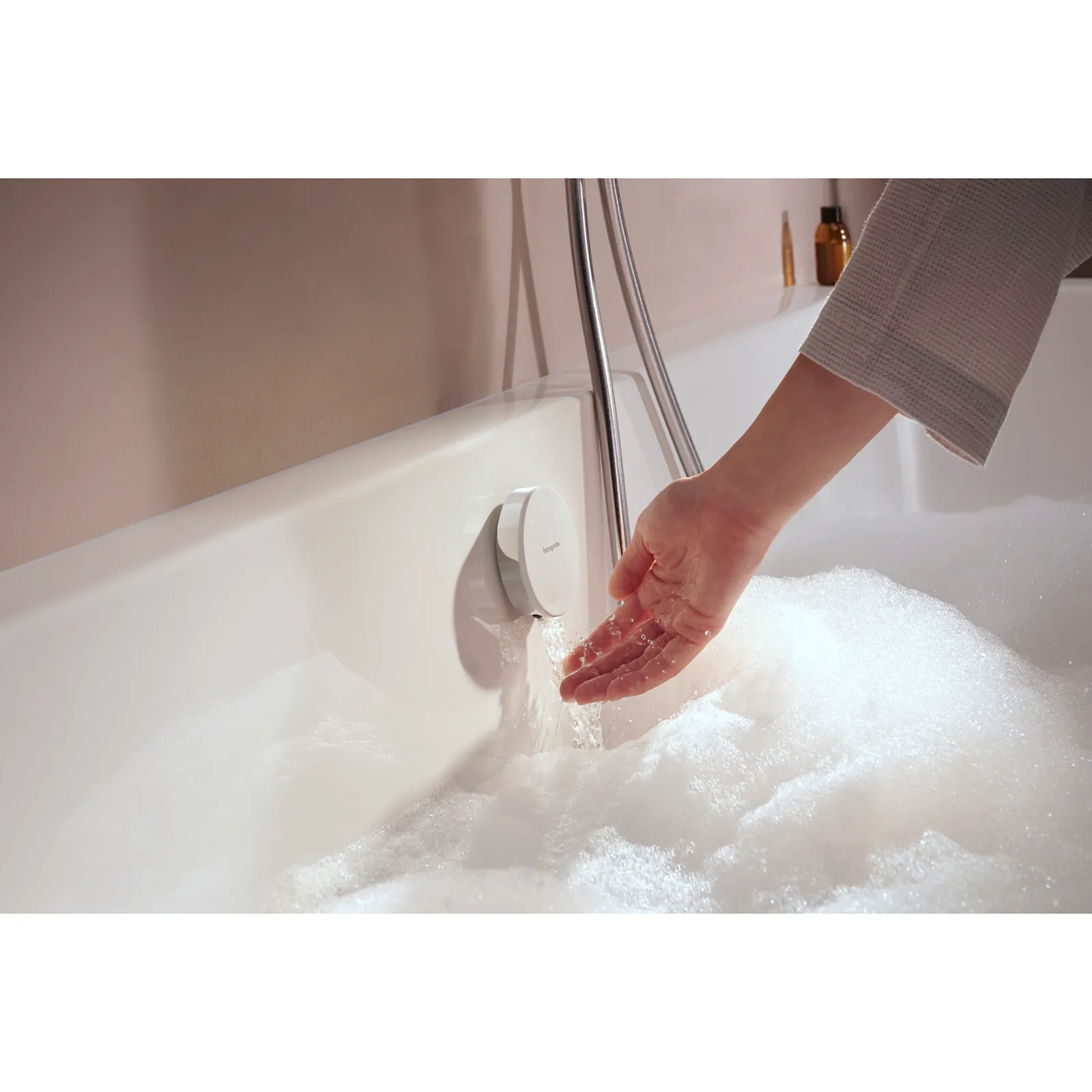 Eine Hand greift nach einem Wasserhahn in einer Badewanne, aus dem Wasser mit Schaum fließt. Die Wanne hat eine weiße, glatte Oberfläche und ist in einer schlicht gestalteten Umgebung.