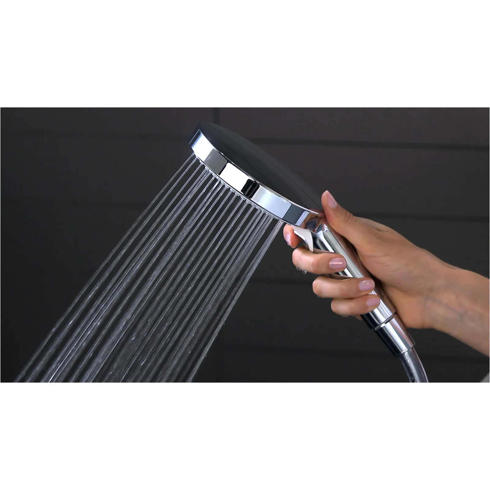 Eine handgehaltene Brause mit glänzendem Chromfinish und einem runden Kopf, aus dem dünne Wasserstrahlen fließen, wird von einer Hand gehalten. Der Hintergrund ist dunkel.